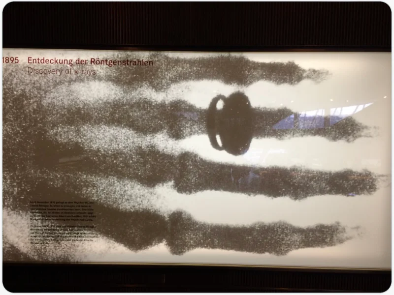 Se ve una foto de la primera radiografía. Es de una mano y se ve un anillo en uno de los dedos. En la parte superior izquierda de la radiografia hay un texto en alemán y el año 1895 en números en color granate.