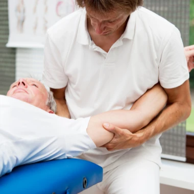Vista de un fisioterapeuta con polo y pantalones blancos sosteniendo un brazo de un paciente sobre una camilla haciendo un ejercicio de recuperación de hombro después de una artroscopia de hombro.