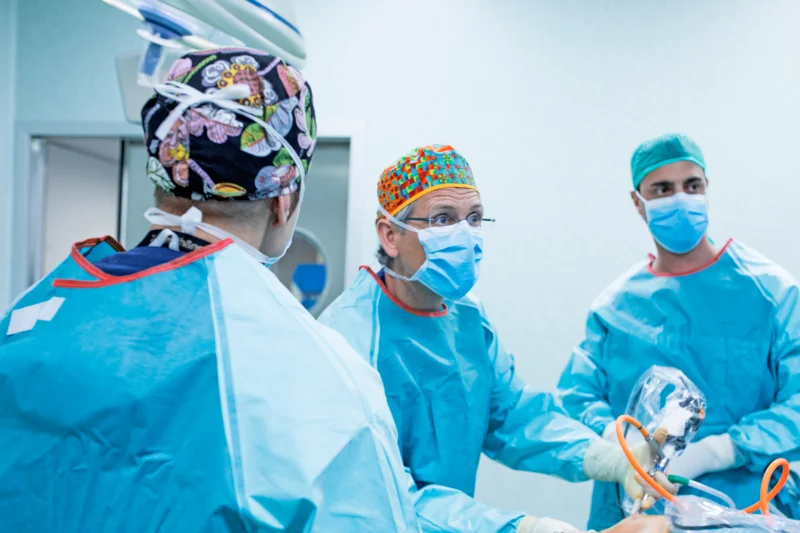 Tres médicos en una sala de operaciones practicando una artroscopia de rodilla. Dos de ellos están mirando hacia la derecha de la foto y el tercero está mirándolos a ellos dando la espalda a la foto.