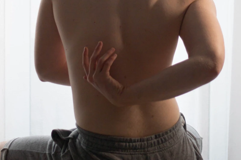 Una persona sentada de espaldas a la cámara, con su parte superior desnuda y con unos pantalones cortos de chándal grises se toca con su mano la parte posterior de la espalda media-baja. Con unas cortinas blancas al fondo.