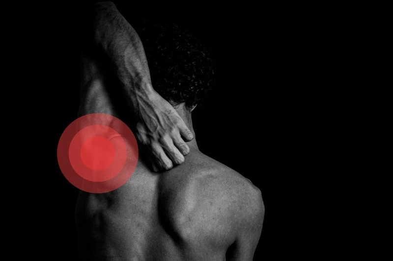 Vista de una foto en blanco y negro, donde se ve un hombre desde la media espalda hasta la cabeza de espaldas a la cámara. Tiene su brazo izquierdo estirado por encima de la cabeza y el codo doblado hacia abajo pasando su antebrazo y mano por detrás de la cabeza cayendo sobre la zona del cuello. Sobre su hombro izquierdo hay unos círculos rojos concéntricos destacando la zona donde se ubican las prótesis de hombro.