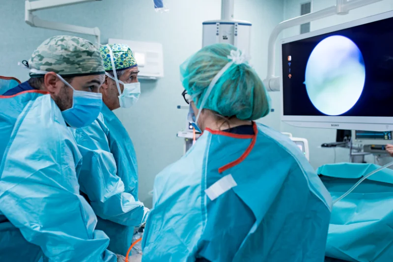 Imagen de tres médicos del Instituto Cugat que están practicando una artroscopia en un quirófano. Un doctor está comentando con una doctora que está de espaldas a la cámara, mientras otro doctor está mirando el monitor de seguimiento de la cámara de la artroscopia que queda en el lado derecho de la foto.