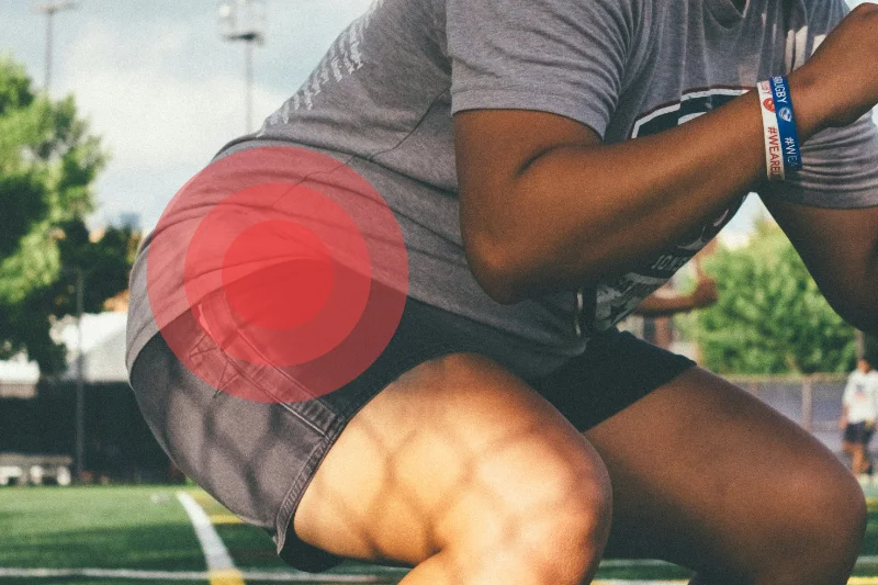 Una persona de la que solo se ve desde la parte inferior de las rodillas hasta pa parte inferior de los hombros está haciendo una sentadilla en lo que parece un campo de fútbol. Lleva pantalones deportivos negros y camiseta gris. Está orientado hacia la parte derecha de la foto y en la zona de la cadera hay unos puntos rojos concéntricos que simbolizan que hay dolor en la zona del labrum de cadera.