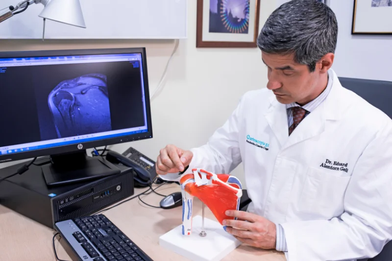 El Dr. Eduard Alentorn sentado en su despacho muestra una maqueta del hombro al tiempo que con su mano derecha describe el síndrome subacromial de hombro. A su derecha queda un ordenador con un monitor que enfoca a la cámara fotográfica y en el que se puede ver una radiografía de hombro.