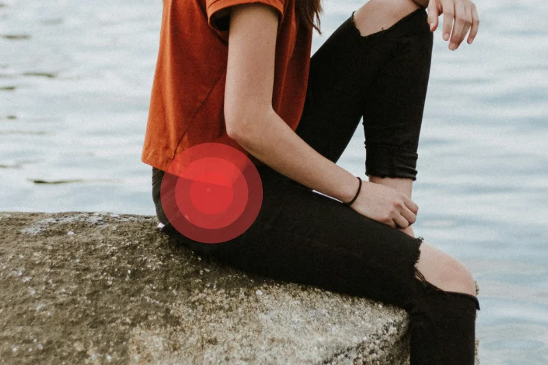 En la foto aparece una chica sentada en una piedra frente a lo que parece un lago. Solo se ve desde el hombro hasta un poco más debajo de la rodilla. Lleva pantalones negros abiertos en ambas rodillas y una camiseta de color naranja oscuro. En la zona de la cadera derecha hay una círculos rojos concéntricos que indican dolor en esa zona