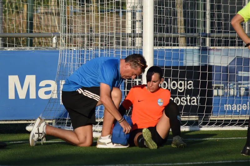 En la foto se ve a un fisioterapeuta arrodillado en el césped de un campo de fútbol junto a una portera que está sentada con una pierna estirada. El fisioterapeuta está revisando la zona de los ligamentos cruzados.