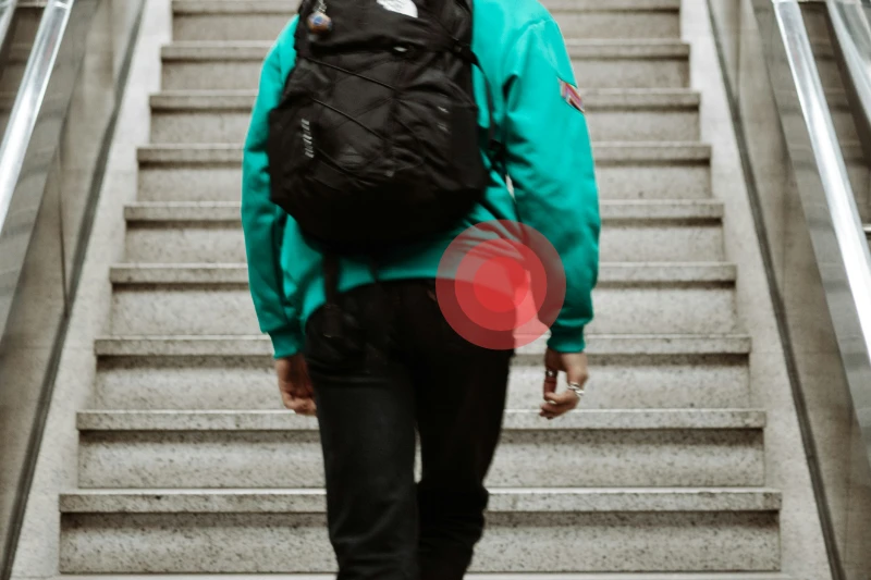 Una persona de la que se ve medio cuerpo, desde las rodillas hasta casi todos los hombros, está subiendo por unas escaleras. Lleva pantalones negros y jersey verde. A la altura de su cadera derecha hay una imagen formada por varios círculos concéntricos en color rojo que indican que hay dolor.