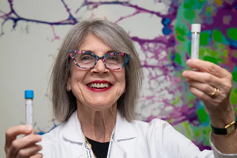 La Dra. Montserrat Garcia Balletbó, con gafas de colores y bata blanca, sonríe mientras sostiene dos tubos de ensayo en sus manos. El fondo muestra una ilustración colorida de conexiones neuronales. Foto propiedad de La Vanguardia.