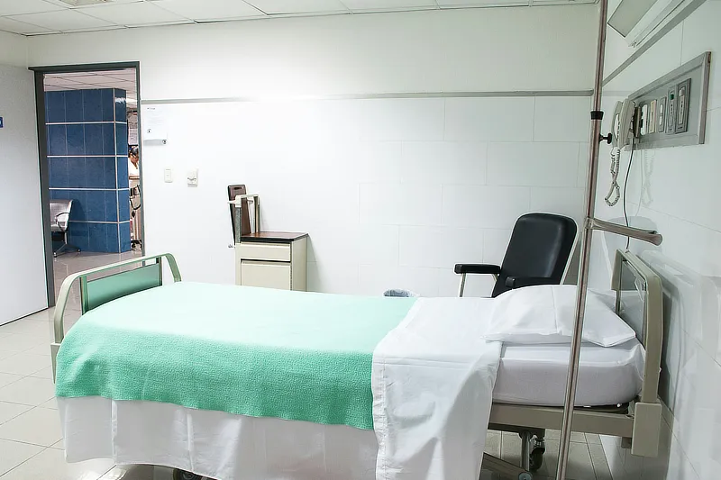 Habitación de hospital vacía con una cama de hospital con sábanas blancas y una manta verde. Al lado de la cama hay una silla negra y una mesita. En la pared hay un panel con tomas eléctricas y un teléfono. La puerta está abierta, mostrando un pasillo azul.