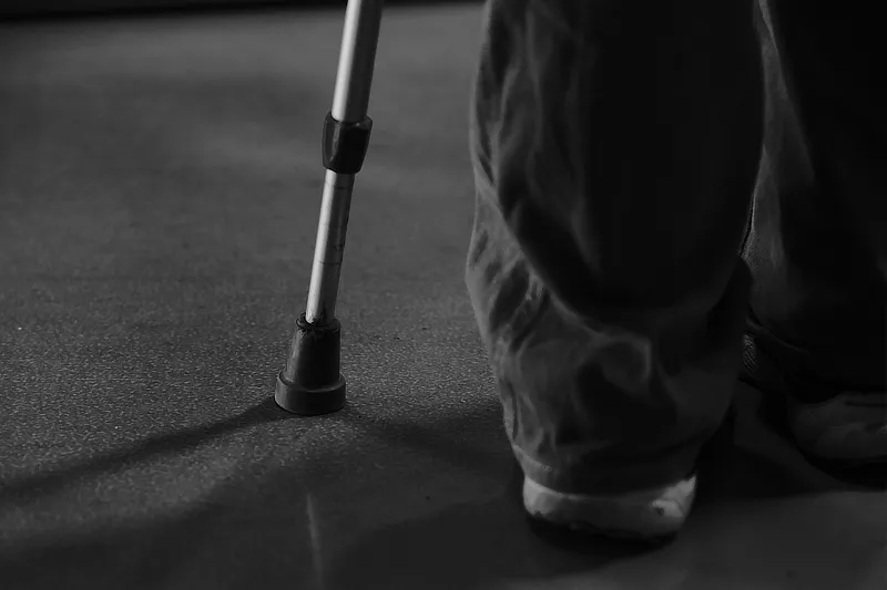 Persona usando una muleta para caminar, ilustrando la necesidad de apoyo debido al desgaste del cartílago de cadera. La imagen en blanco y negro muestra una vista de cerca de la muleta y los pies de la persona, destacando el uso de equipo de asistencia para la movilidad.