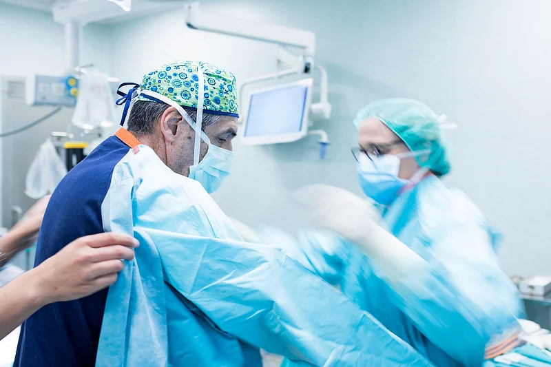 Cirujano en el quirófano siendo asistido por personal médico para colocarse la bata esterilizada antes de realizar una cirugía artroscópica de cadera. Monitor y equipo médico visible en el fondo.