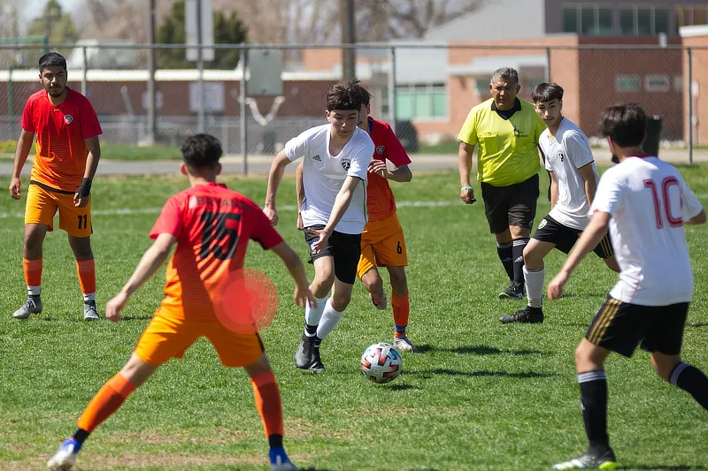 Un grupo de jóvenes jugando un partido de fútbol en un campo de césped. Un jugador del equipo con camisetas blancas dribla el balón, rodeado por jugadores del equipo con camisetas naranjas. Un círculo rojo destaca la cadera de un jugador, indicando dolor o una lesión en esa área. Un árbitro, vestido de amarillo, observa de cerca la acción mientras otros jugadores están atentos. En el fondo se pueden ver edificios y una valla.