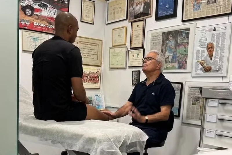 El Dr. Ramón Cugat trata a Fernandinho en su clínica de Barcelona: El Dr. Cugat evalúa la rodilla de Fernandinho, sentado en una camilla, rodeado de diplomas y fotos que destacan su carrera. La imagen refleja la experiencia médica y el enfoque personalizado en la recuperación de lesiones deportivas.