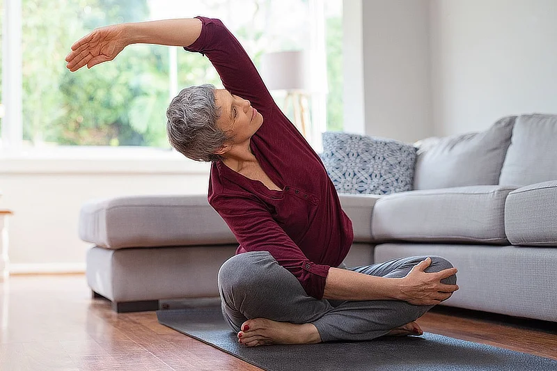 Mujer mayor sentada en una esterilla de yoga en su sala de estar, realizando un estiramiento lateral con el brazo izquierdo elevado. Lleva camiseta burdeos y pantalones grises, en un entorno hogareño y luminoso.