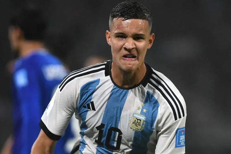 Valentín Carboni, futbolista argentino, aparece en la imagen vistiendo la camiseta de la selección argentina. Lleva el dorsal número 10 y se le ve haciendo una expresión de esfuerzo o intensidad durante el partido. De fondo, se observan otros jugadores desenfocados, destacando la figura de Carboni en primer plano.