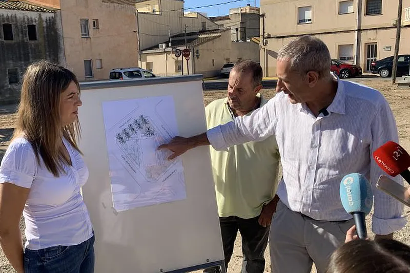Tres personas, dos hombres y una mujer, observan un plano arquitectónico colocado en un caballete al aire libre. Uno de los hombres señala detalles del plano mientras explica el proyecto a los otros, quienes prestan atención. Una periodista sostiene un micrófono que apunta hacia los participantes. Al fondo, se ven edificios residenciales y vehículos estacionados. Foto: L’Aldea (laldea.cat).