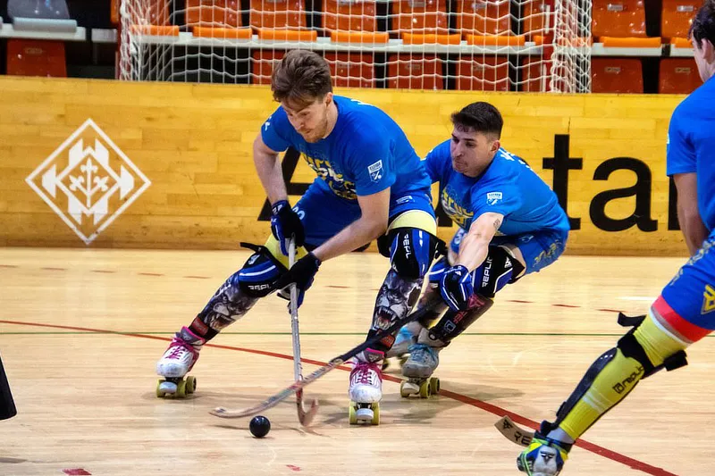 Dos jugadores de hockey patines, con camisetas azules y equipación de protecciones, luchan por el control de una pelota en la pista. El jugador al frente parece tener el control mientras el otro intenta alcanzarla desde atrás. Al fondo, se ve una portería y las gradas vacías del pabellón deportivo.