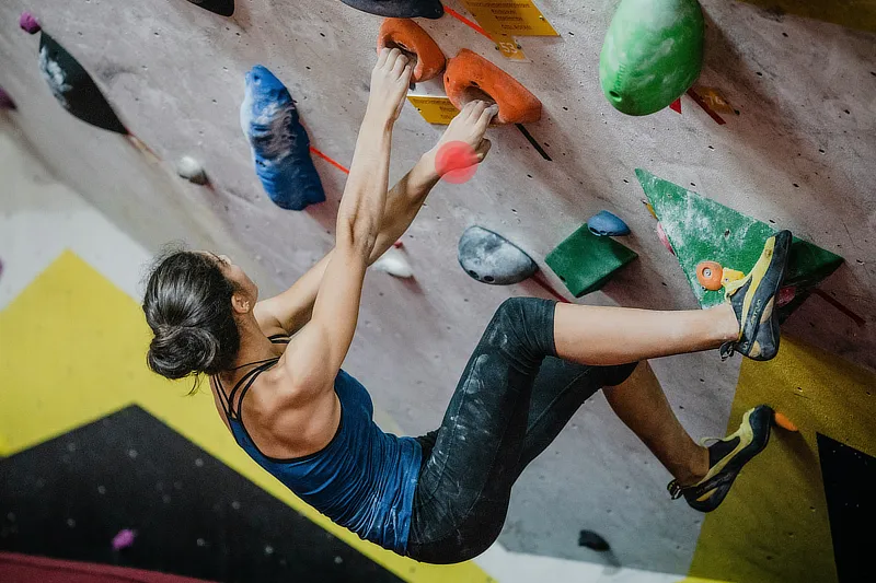 Una mujer escaladora en un rocódromo realizando una postura desafiante, con énfasis en su mano derecha mientras agarra una presa con fuerza. La imagen destaca el esfuerzo y el potencial riesgo de lesiones en el pulgar y muñeca debido al sobreuso repetitivo.