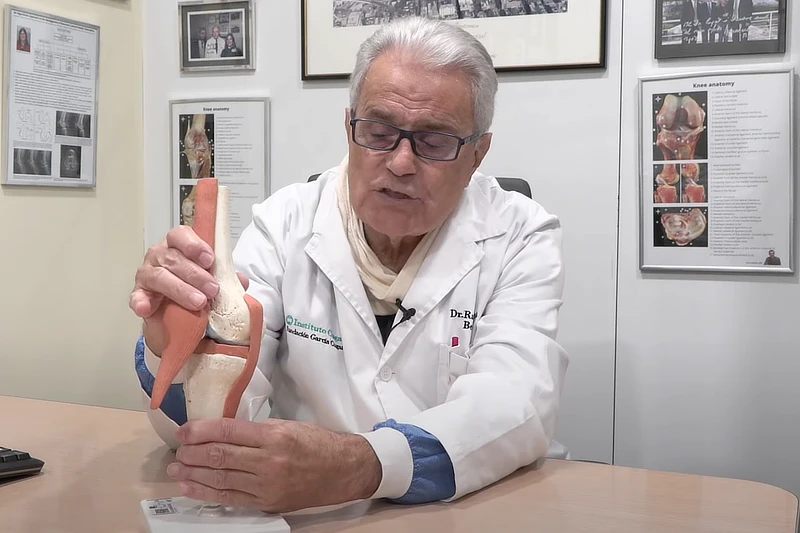 El Dr. Ramón Cugat, con bata médica, sostiene un modelo anatómico de rodilla mientras explica su funcionamiento en su consulta, rodeado de cuadros, diagramas médicos y material informativo.