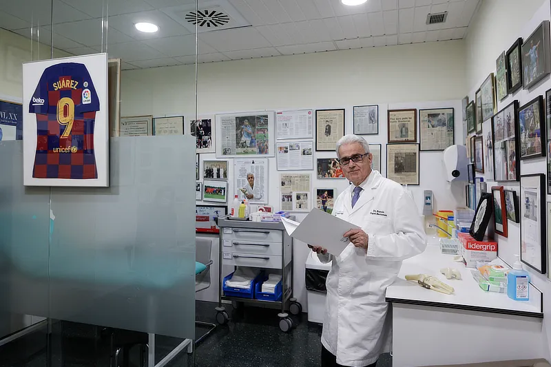 El Dr. Ramón Cugat en su consulta, sosteniendo documentos, con fotografías y artículos enmarcados en la pared. A la izquierda, una camiseta firmada de Luis Suárez.