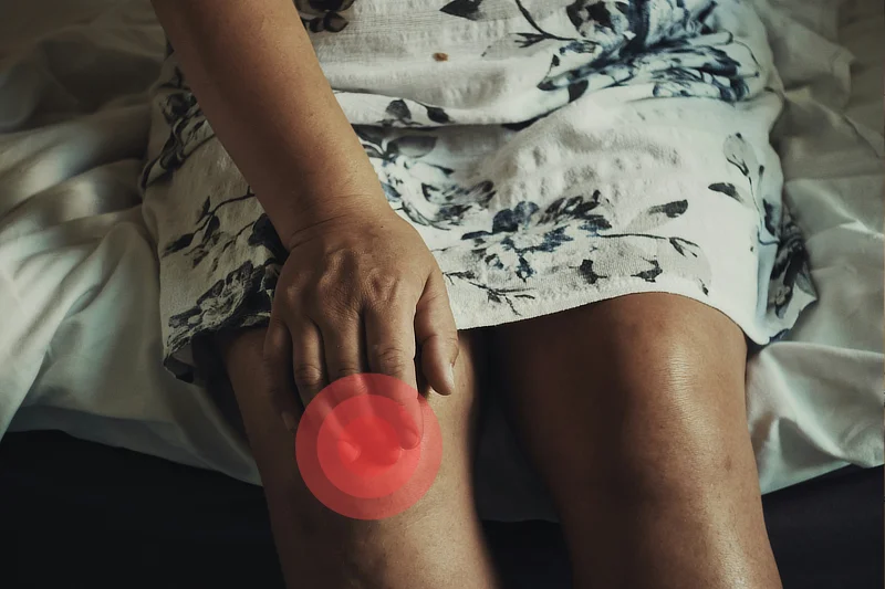 Una mujer sentada sobre una cama, con un vestido claro de flores, sostiene su rodilla derecha con gesto de dolor. La imagen resalta la articulación con un círculo rojo para indicar la zona afectada por posible artrosis de rodilla.