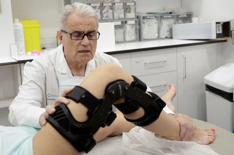 El Dr. Ramon Cugat examina la rodilla de una paciente equipada con una rodillera ortopédica negra mientras realiza una evaluación clínica en la consulta.