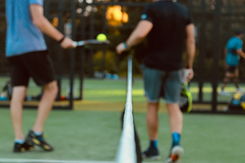 Dos jugadores de pádel en una pista al aire libre, vistos de espaldas y con la red en primer plano, listos para comenzar un partido.