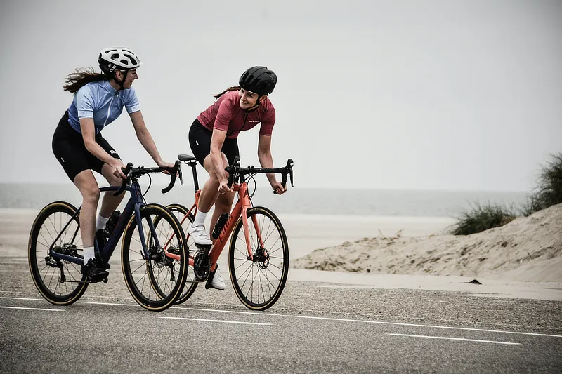 Dos ciclistas mujeres pedaleando en carretera cerca de la playa, con cascos y ropa deportiva, disfrutando de la actividad física al aire libre.
