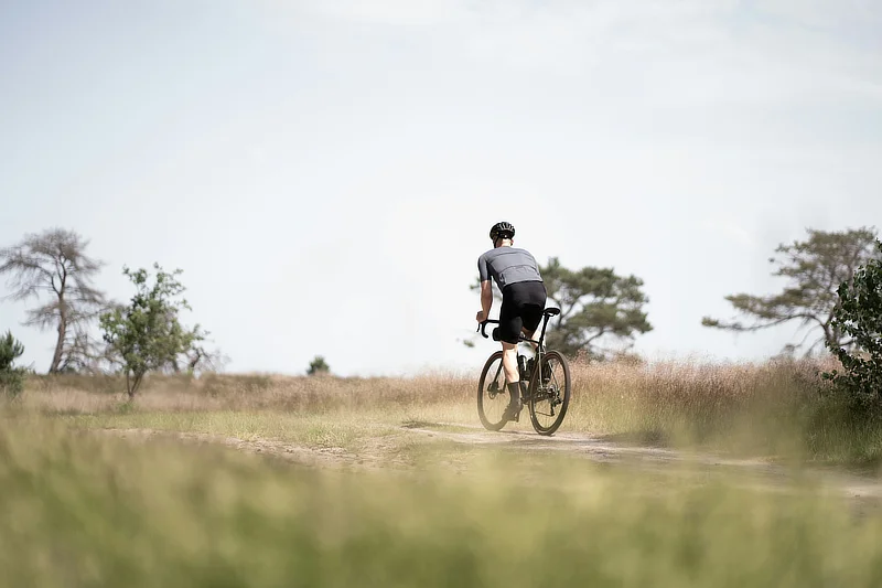 Ciclista hombre visto desde atrás pedaleando solo por un camino en el campo, con vegetación y árboles alrededor en un entorno natural.