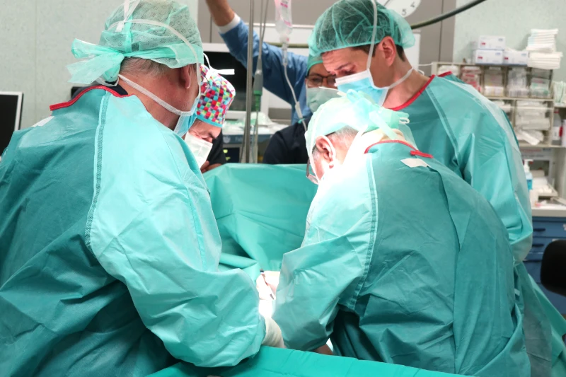 Equipo de cirujanos con bata estéril y mascarillas realizando una osteotomía valguizante de tibia en el quirófano, rodeados de instrumental médico y luz quirúrgica enfocada en la rodilla del paciente