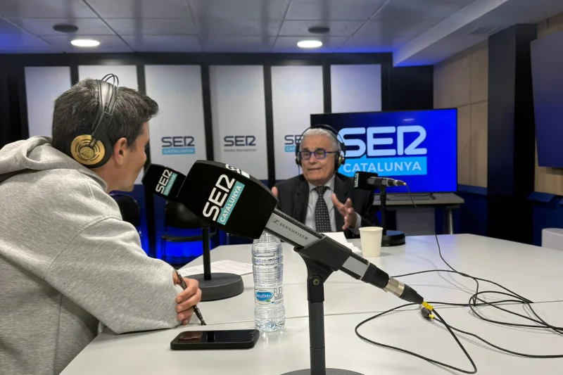 El Dr. Ramón Cugat conversa con el locutor de SER Catalunya en el estudio de radio, gesticulando mientras responde, con micrófonos y pantallas del programa visibles al fondo.