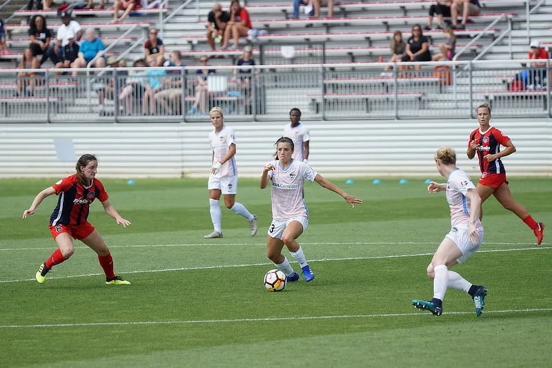 Partido de fútbol femenino en campo de césped natural, con varias jugadoras en acción durante una jugada ofensiva; una de ellas conduce el balón mientras otras se preparan para defender o recibir el pase, con gradas llenas de espectadores al fondo.