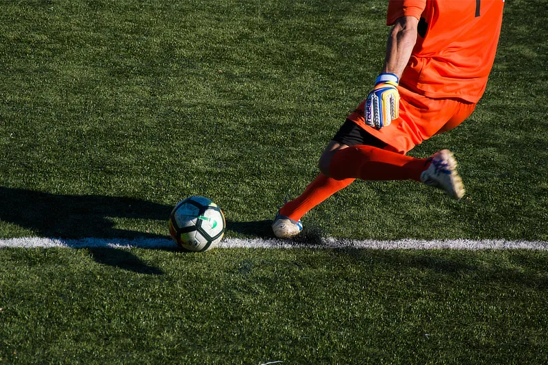 Portero de fútbol realiza un potente chut con la pierna derecha, captado justo en el momento del impacto con el balón sobre el césped artificial.