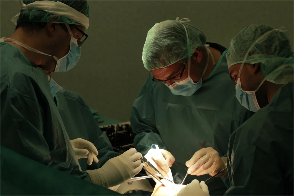 Three surgeons in green scrubs, surgical masks, caps, and protective glasses working with deep focus under an intense focal light inside an operating room during a surgery.