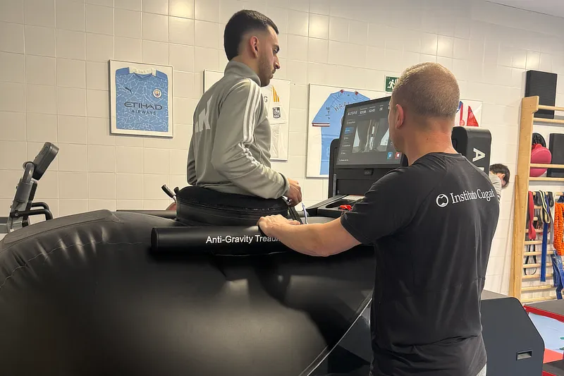 Fotografía de perfil de un hombre caminando sobre la cinta Alter G en una sala de rehabilitación con ventanales al fondo. El paciente viste ropa deportiva oscura y está rodeado por la cámara de aire blanca de la máquina, que llega hasta su cintura.