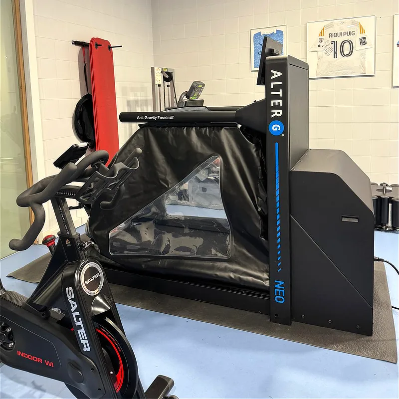 Fotografía de la cinta de correr antigravedad Alter G modelo NEO en una sala de rehabilitación de instituto Cugat en Barcelona. En primer plano aparece una bicicleta estática Salter y al fondo, en la pared, una camiseta enmarcada del jugador Riqui Puig. Se aprecia el lateral de la máquina con su estructura negra y la cámara de aire con ventana transparente.