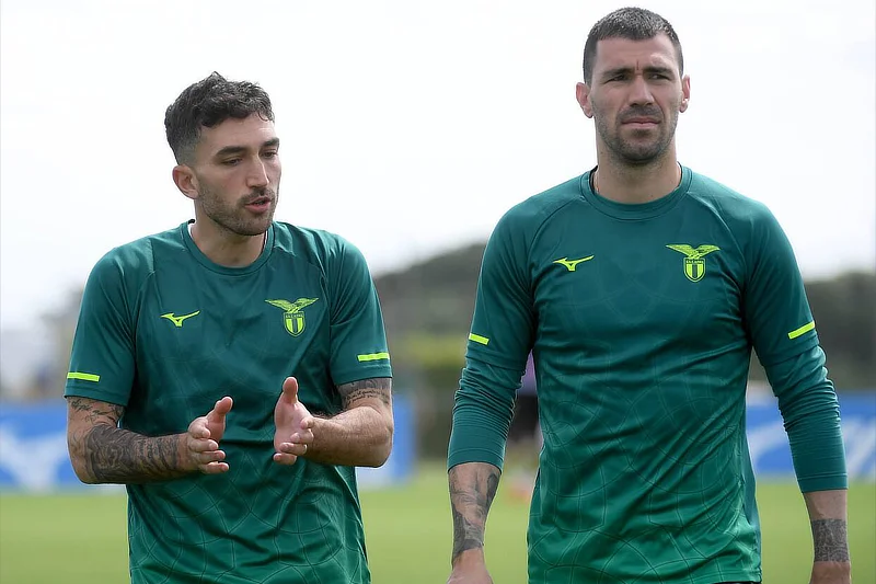 Dos jugadores de fútbol con camisetas verdes de entrenamiento de la Lazio caminando sobre el césped; el de la izquierda realiza un gesto explicativo con las manos mientras habla. Foto OneFootball.