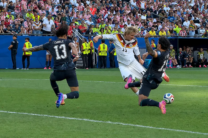 Jugador de la selección alemana conduce el balón mientras dos futbolistas rivales intentan frenar su avance con una entrada durante un partido del Mundial. La acción se desarrolla cerca del área, con las gradas llenas de aficionados al fondo.