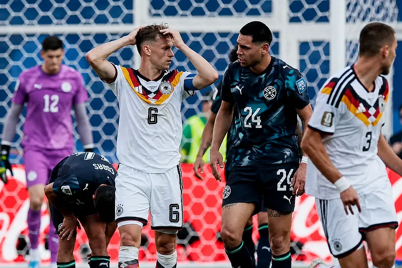 Jugadores de Alemania y su selección rival durante una pausa del partido tras una acción de juego. Un futbolista alemán se lleva las manos a la cabeza mientras otros jugadores permanecen cerca del área, con la portería y el guardameta al fondo.