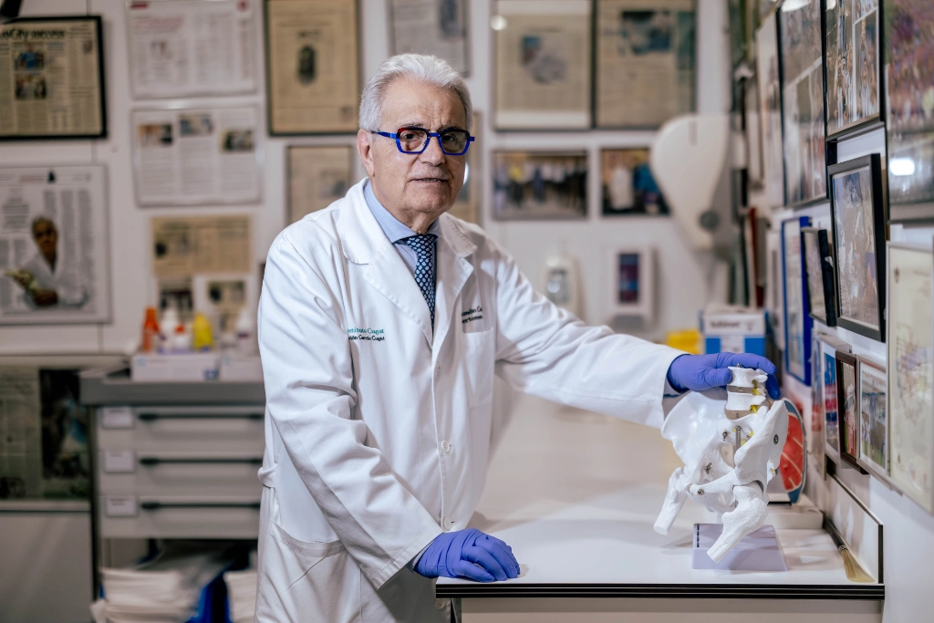 Dr Ramón Cugat wearing a white coat and blue gloves in his consultation room, with his hand resting on an anatomical model of the pelvis and spine.