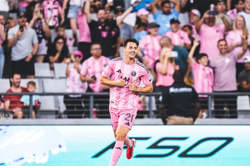 Jugador Tadeo Allende del Inter Miami con uniforme rosa sonríe y corre por el campo celebrando un gol, con la afición en la grada de fondo vistiendo camisetas del mismo color.