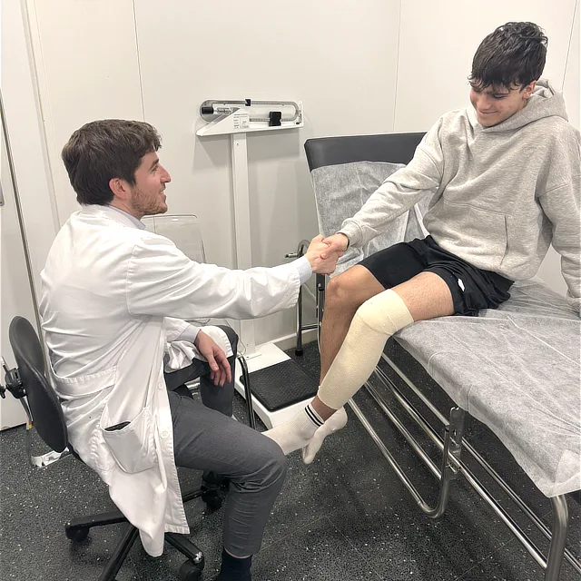 Unidad de fracturas 3 Que tratamientos realizamos Doctor saludando a un paciente joven con la pierna vendada sentado en una camilla de consulta.