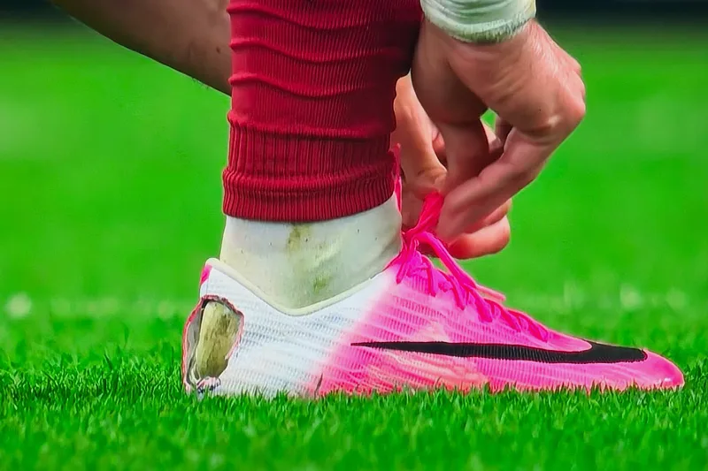 Primer plano de un futbolista atándose los cordones de una bota rosa y blanca que presenta un agujero recortado a mano en la parte posterior del talón.