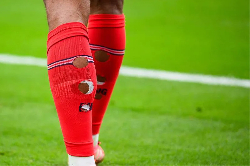 Detalle de los gemelos de un jugador de fútbol de espaldas mostrando tres agujeros redondos recortados a mano en sus medias rojas de compresión.