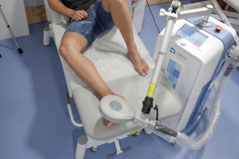 Paciente en camilla recibiendo tratamiento con tecnología médica en la zona trasera y media del pie durante la rehabilitación de una artroscopia de tobillo.